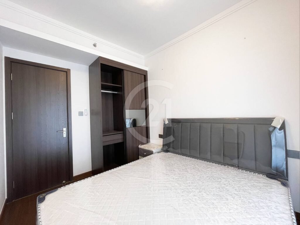 A minimal bedroom with a dark wooden door, an open wardrobe, a nightstand, and a bed frame with a mattress covered in plastic. The walls are white and the room is well-lit.