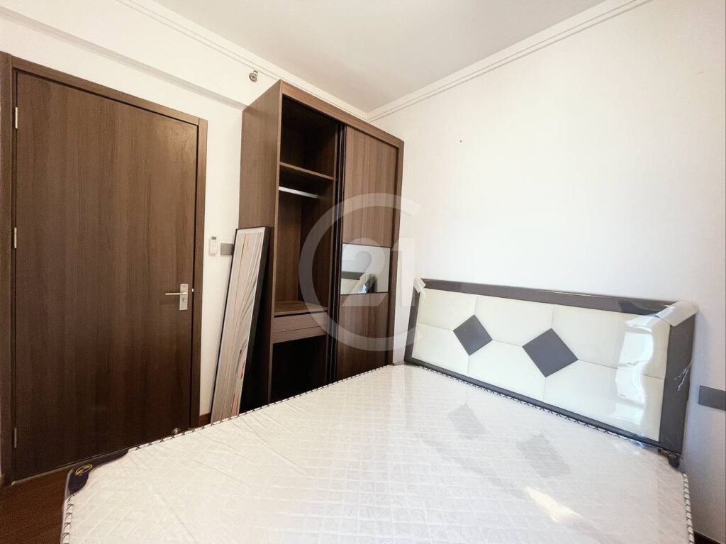 A small bedroom with a wooden door, an open wardrobe, a large mirror propped against the wall, and a bed with a white and black geometric headboard and a mattress covered in plastic.