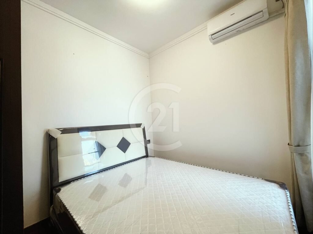 A small, minimalist bedroom with a bed featuring a white quilted mattress and a two-tone headboard, cream walls, an air conditioner above, and a window with curtains on the right.