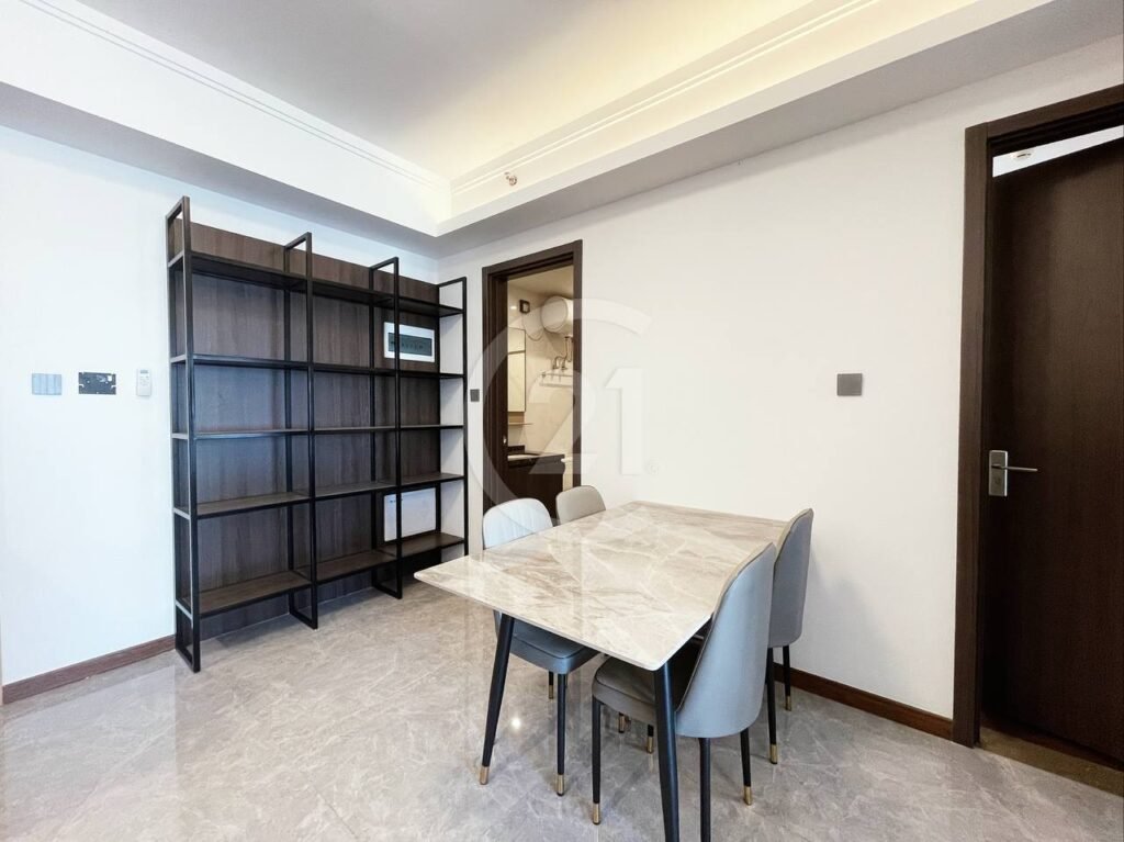Modern dining area with a marble table and four gray chairs, set on a light tiled floor. A dark wood open shelving unit stands against the wall, and a dark-framed door is visible on the right. The space is bright and minimalistic.