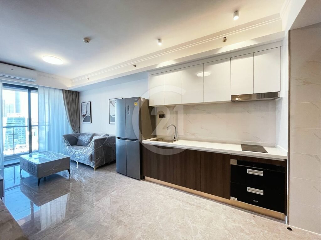 Modern apartment with a bright open-plan kitchen and living area, featuring white and dark brown cabinets, stainless steel fridge, covered sofa, coffee table, and floor-to-ceiling windows overlooking a cityscape.