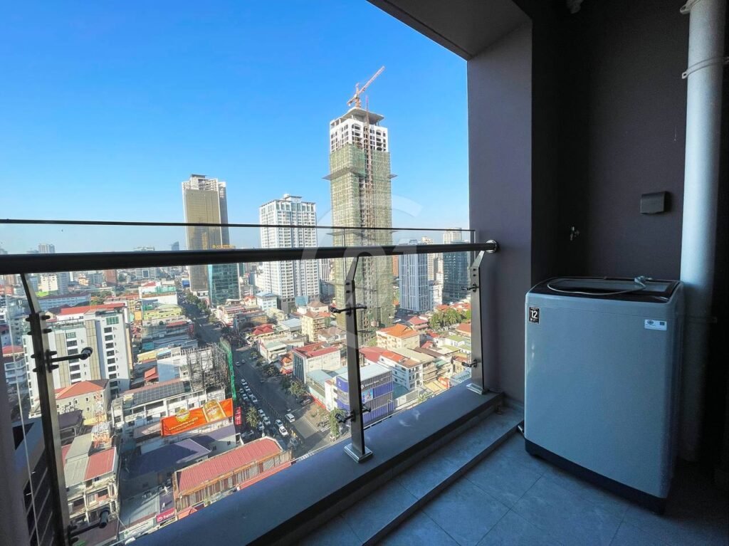 A city view from a high balcony with glass railing, overlooking tall buildings and construction. A washing machine is placed in the corner of the balcony under clear blue sky.