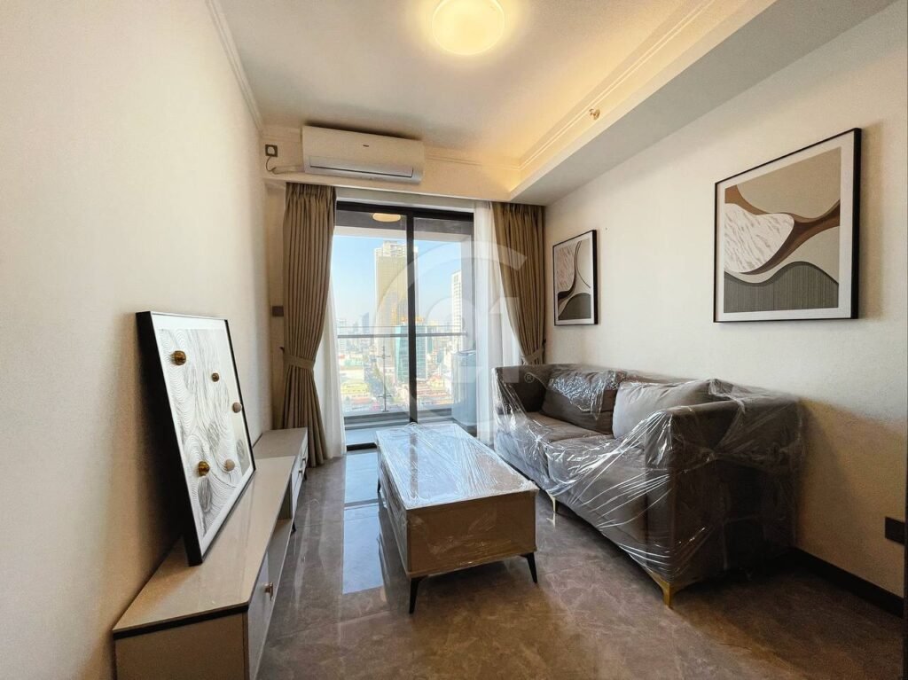 A modern living room with a gray sofa and coffee table covered in plastic, wall art, a cabinet, and floor-to-ceiling windows offering a city view with tall buildings in the background.