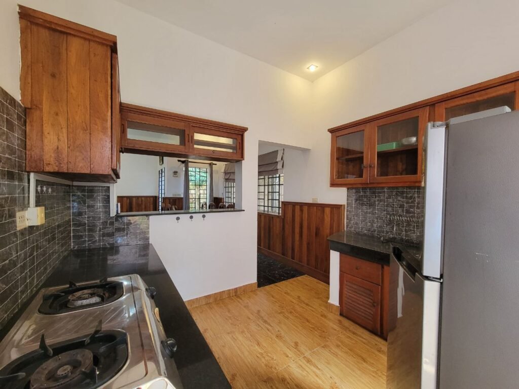 A modern kitchen with wooden cabinets, black countertops, a gas stove, stainless steel refrigerator, tiled backsplash, and light wood flooring. A serving window opens to an adjacent room with natural light.