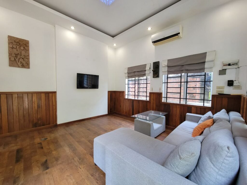 A modern living room with wooden floors and paneling, a light gray sectional sofa with an orange pillow, a glass coffee table, wall-mounted TV, windows with gray Roman shades, and an air conditioner above.