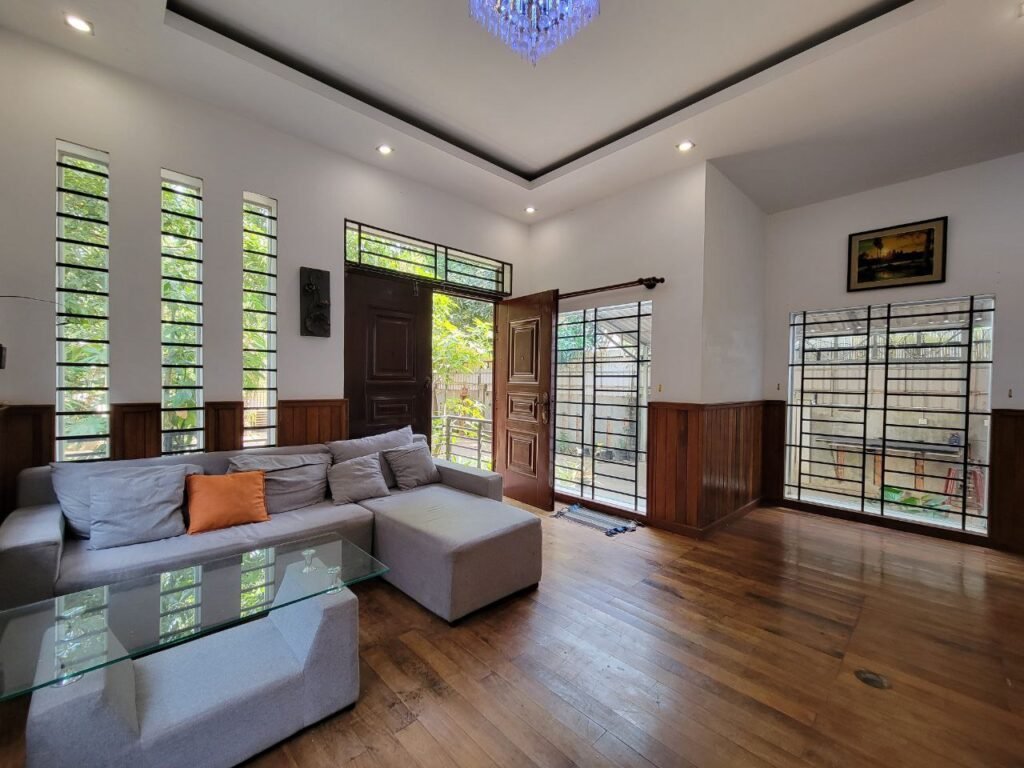 A modern living room with wooden floors, a light gray sectional sofa with an orange pillow, a glass coffee table, large windows with metal grills, and a wooden door open to a sunny garden outside.
