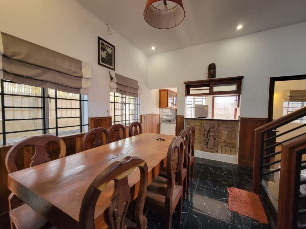 A dining room with a wooden table and eight chairs, black tile floor, large windows with gray blinds, and a view into a kitchen. There is a wall-mounted artwork and a red rug near the staircase.