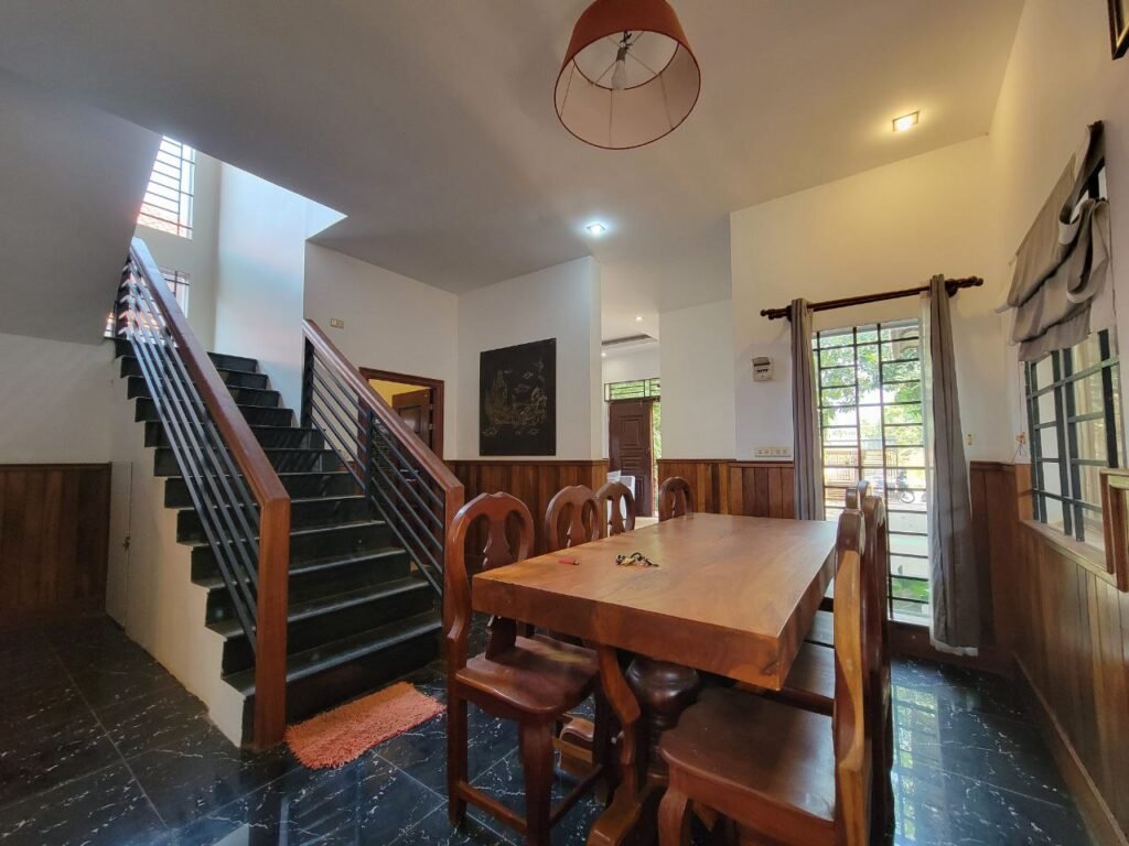 A dining area with a wooden table and chairs, black tiled floor, a staircase with dark steps, wood paneling on the walls, large windows, and natural light coming through open doors and windows.