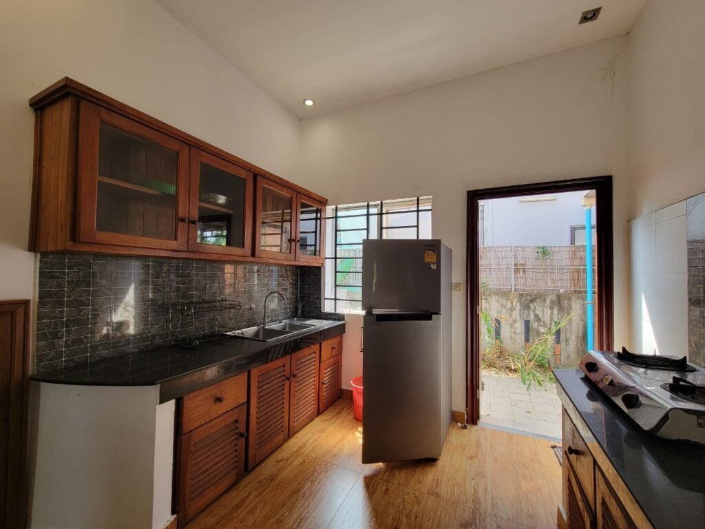 A small kitchen with wooden cabinets, a black countertop, a refrigerator, a sink under a window, and a gas stove. Sunlight enters through an open door leading to a small outdoor area.