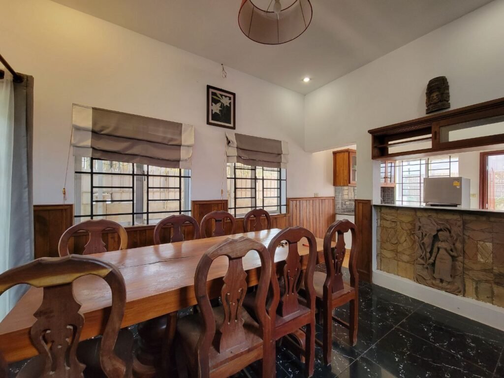 A dining room with a wooden table and eight chairs, two windows with gray Roman shades, and a partial view of a kitchen through a serving hatch with stone detailing and an artistic carving below.