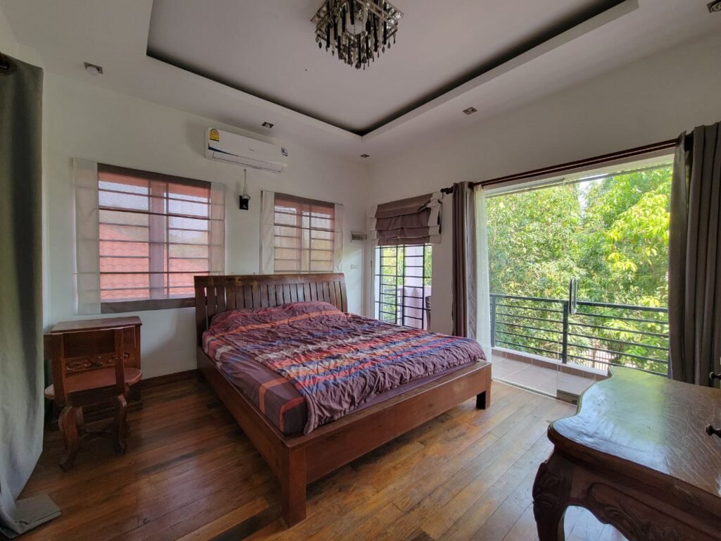 A bright bedroom with a wooden floor and bed, bedside tables, a chandelier, large windows, and a door leading to a balcony with green trees outside.