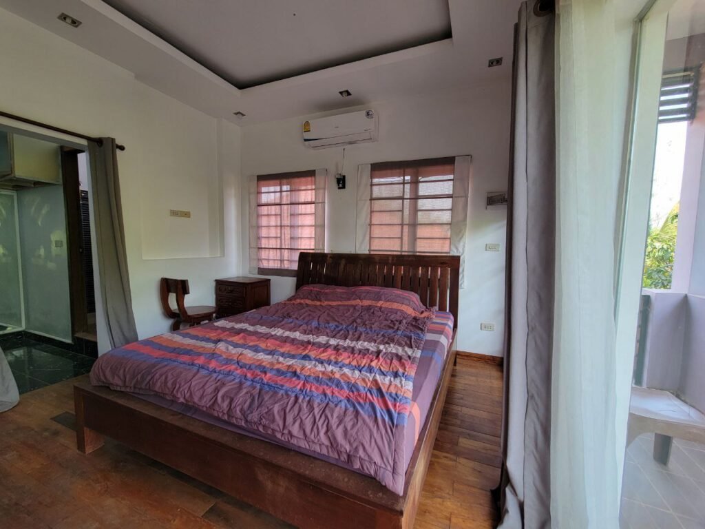 A bright bedroom with a wooden bed, striped comforter, wooden chair, small desk, air conditioning unit above the window, and large glass doors leading to a balcony. Light fills the room from outside.