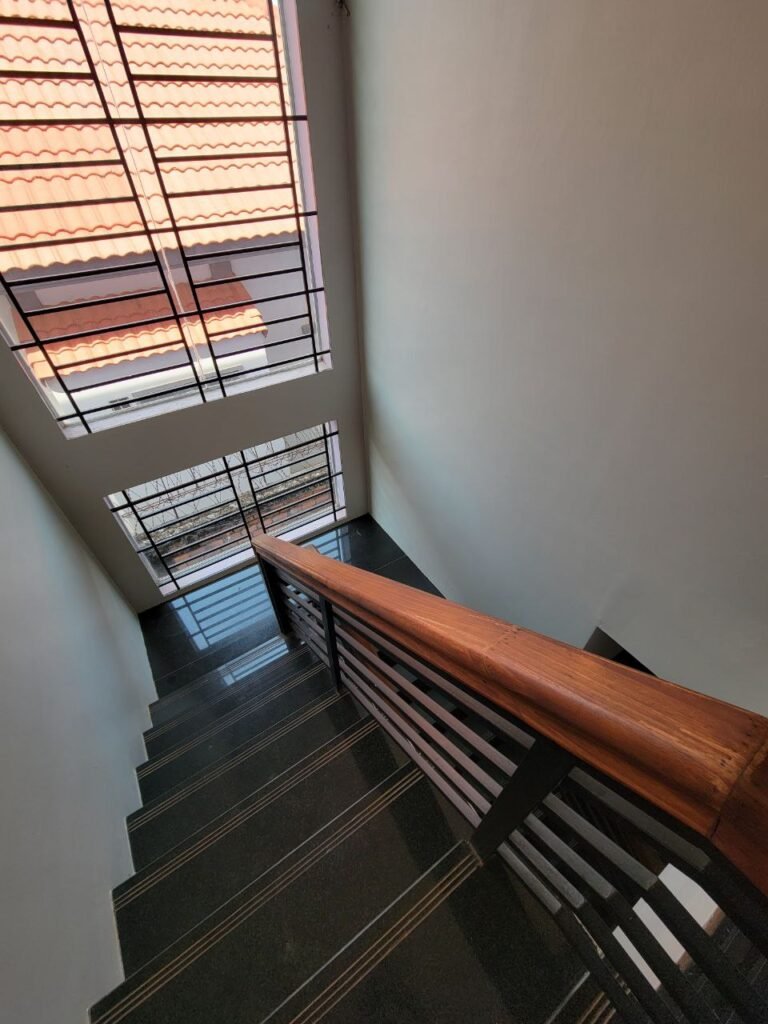 View down a staircase with black steps and a wooden handrail, leading to a glass door. Large windows above the door reveal a tiled roof and let in natural light, brightening the narrow stairwell.
