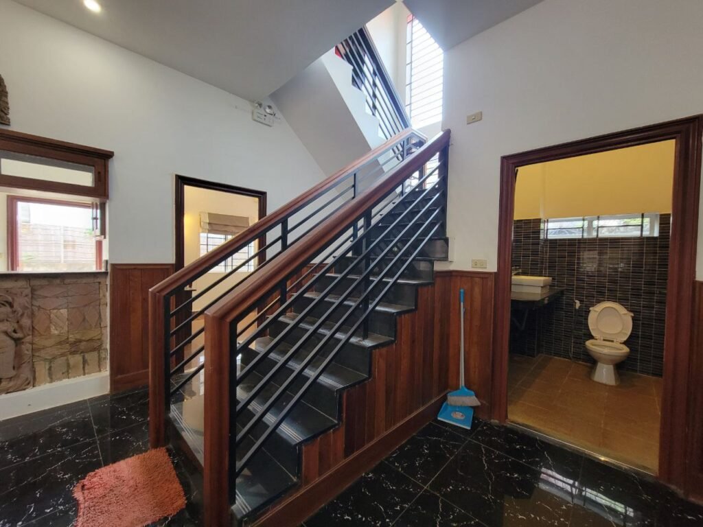 Interior of a house with a staircase in the center, a door to a bathroom with a toilet visible on the right, a red rug on a black marble floor, and a blue broom leaning against the stairs.