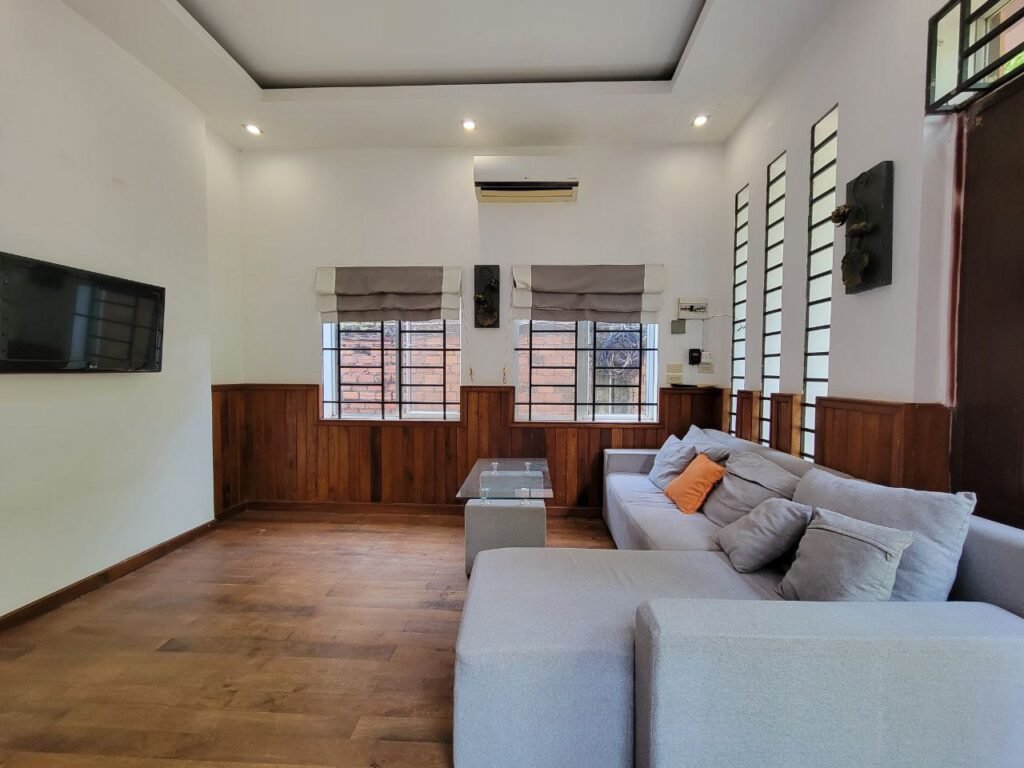 A modern living room with wood flooring, white walls, a gray sectional sofa with orange and gray pillows, a glass coffee table, wall-mounted TV, and large windows with wooden paneling.