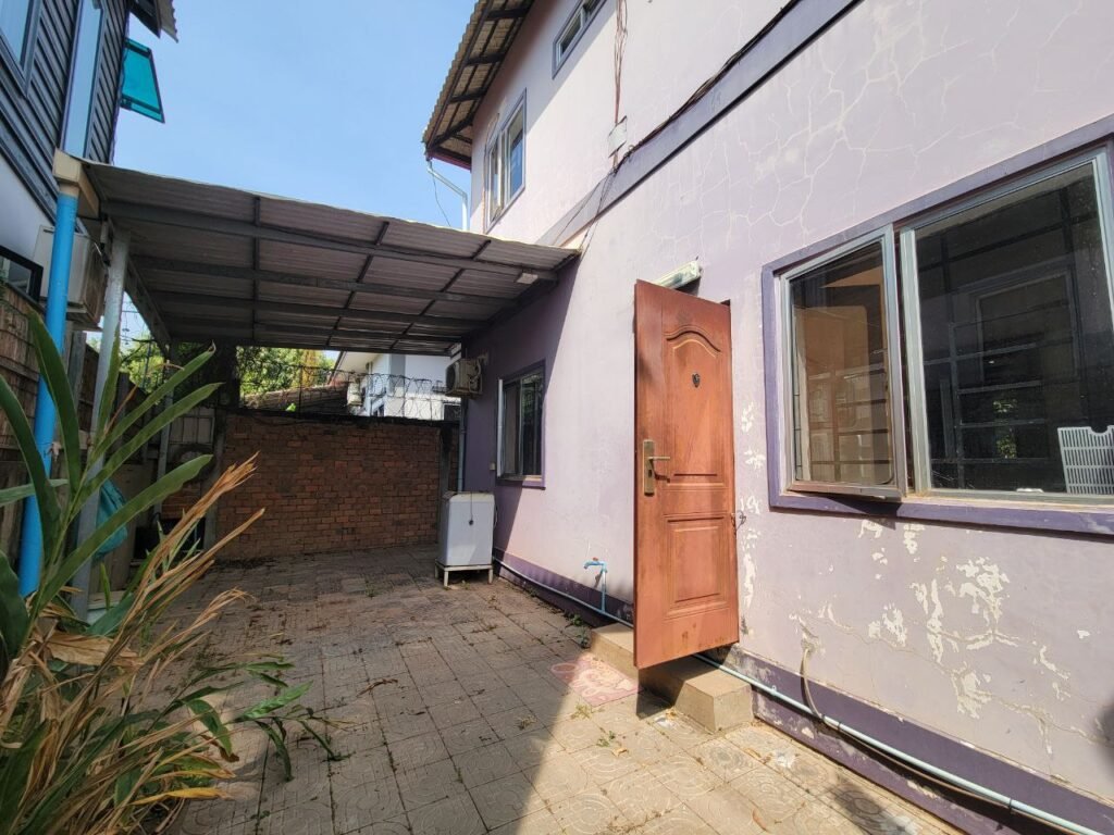 A sunlit outdoor patio with a purple two-story building, open brown door, window, and a covered area with a white appliance. The ground is paved with tiles and bordered by plants on the left.