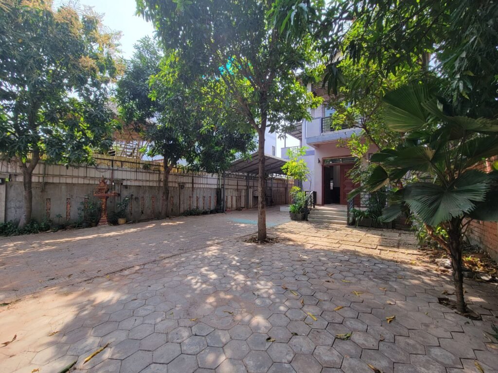 A spacious courtyard with hexagonal paving stones, surrounded by trees and plants, leads up to a house with a covered area and shaded entrance under a clear sky.