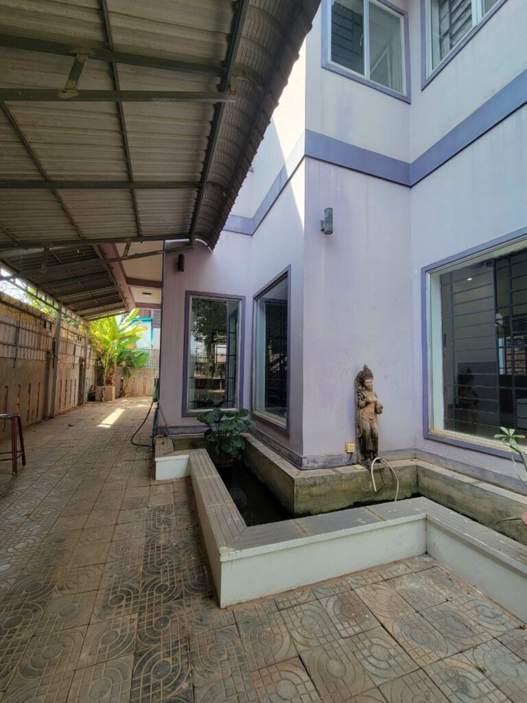 A side view of a house with purple walls, large windows, a covered walkway, and a small outdoor pond featuring a statue fountain. Sunlight casts shadows on the tiled floor, and green plants are visible in the corner.