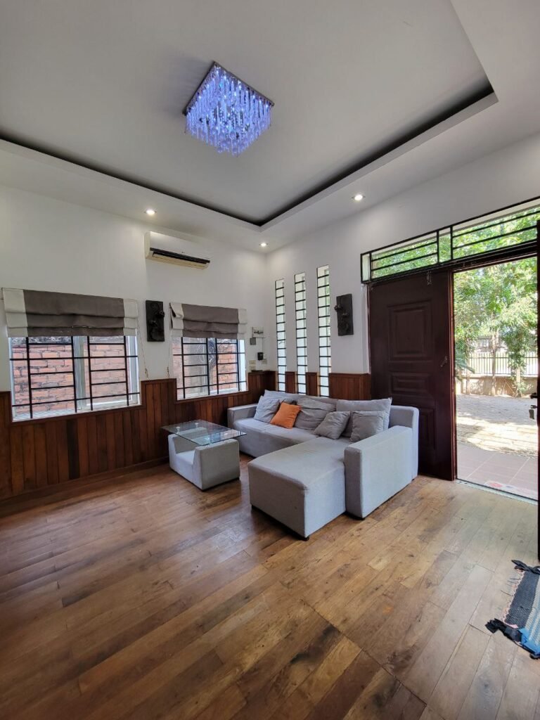 A cozy living room with wooden floors, white walls, a gray sectional sofa with orange and gray cushions, a glass coffee table, and a chandelier. Large windows and an open door reveal greenery outside.