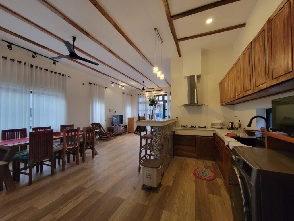 A spacious, modern open-plan kitchen and dining area with wooden furniture, tiled flooring, large windows with white curtains, ceiling fan, and pendant lights. The kitchen has wood cabinets and a countertop with patterned tiles.