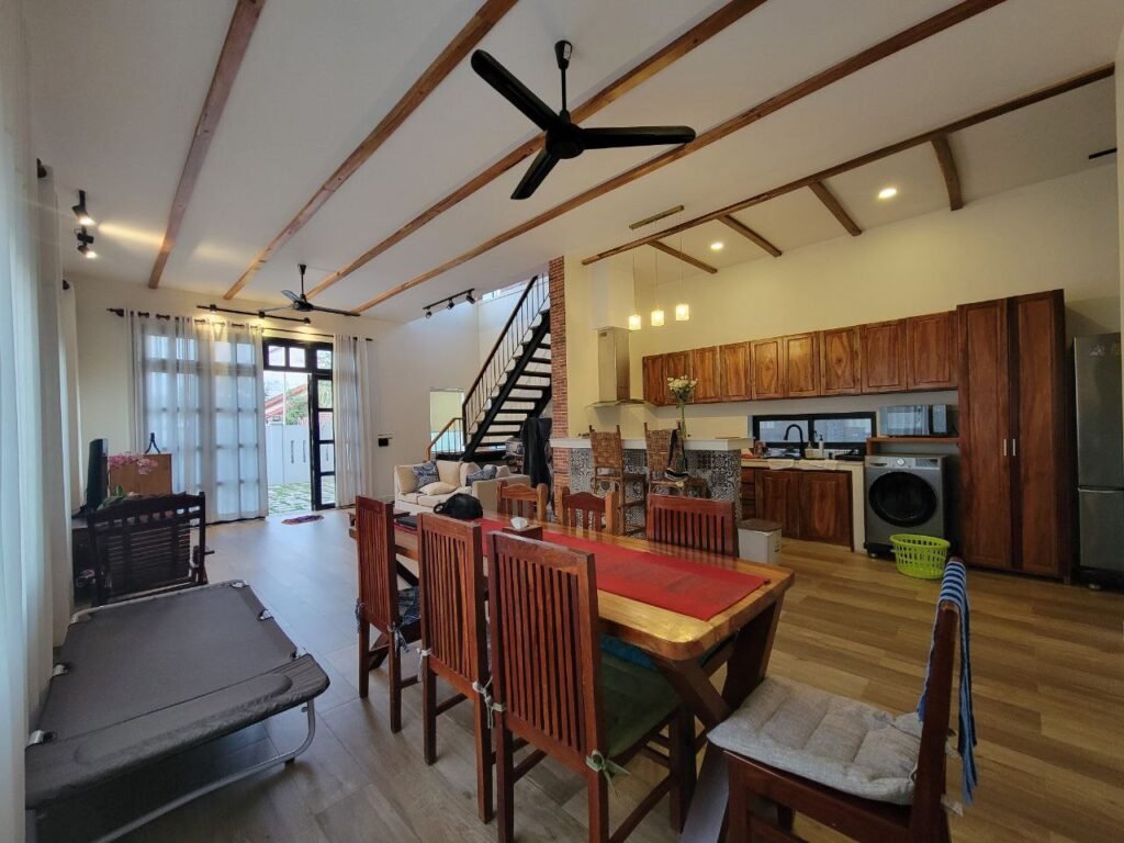 A spacious, modern open-plan living area with wooden floors, a dining table with chairs, a kitchen with wooden cabinets, a staircase, large windows, ceiling fans, and natural light filling the room.
