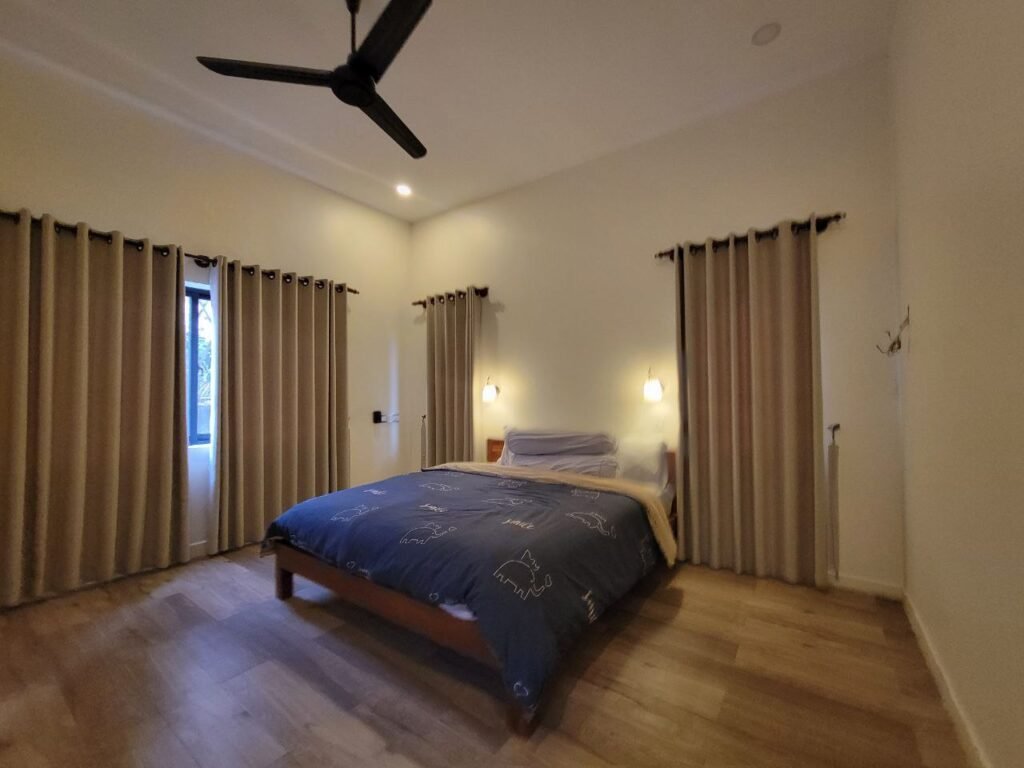 A cozy bedroom with a double bed covered in a dark blue duvet, two bedside lights, light beige curtains on three windows, wooden flooring, and a ceiling fan. The room is simply furnished and well-lit.