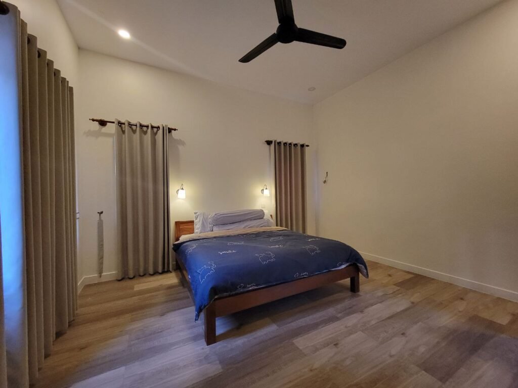 A minimalist bedroom with light-colored walls, wooden flooring, a double bed with blue bedding, wall-mounted lamps, beige curtains, and a black ceiling fan. The room appears tidy and spacious.