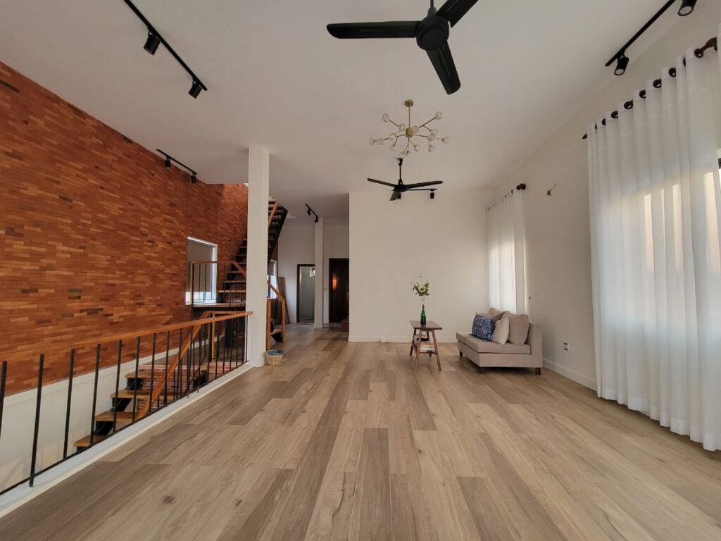Spacious modern living room with light wood flooring, a brick accent wall, black ceiling fans, white curtains, a sofa with cushions, a small table with a vase, and a staircase with black railings.
