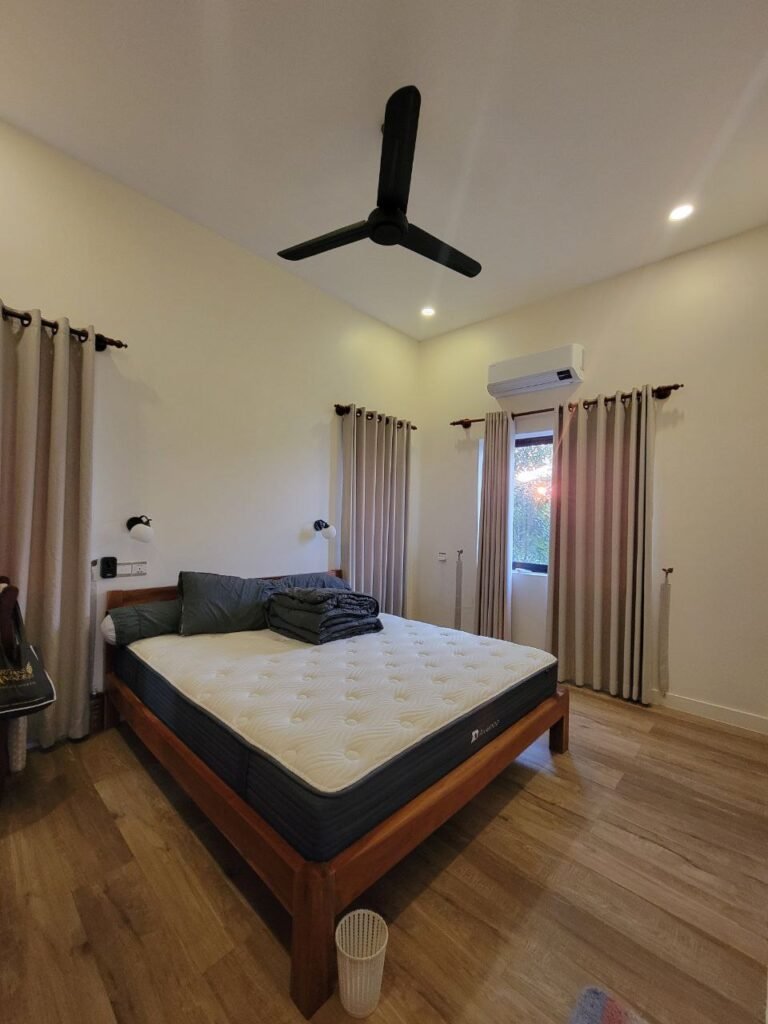A neatly made bed with a mattress sits in a modern, minimalist bedroom with wooden flooring, beige curtains, a ceiling fan, wall-mounted reading lights, and a window showing greenery outside.