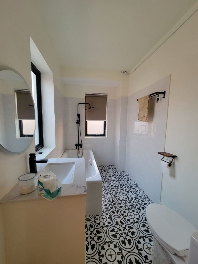 A modern bathroom with patterned black-and-white floor tiles, a bathtub with a black shower fixture, a window with a beige blind, a round mirror, and a toilet next to a towel hanging on the wall.