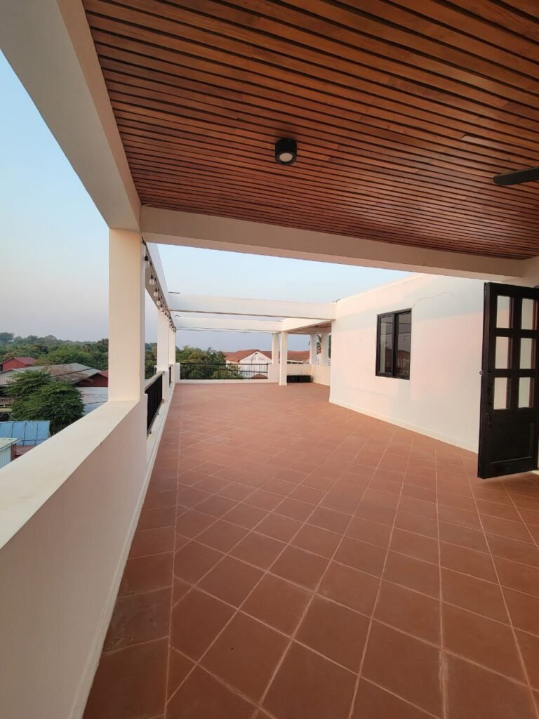 A spacious balcony with terracotta tiles, white walls, and a wooden ceiling overlooks rooftops and greenery at sunset. Glass doors and large windows are visible on the building wall.