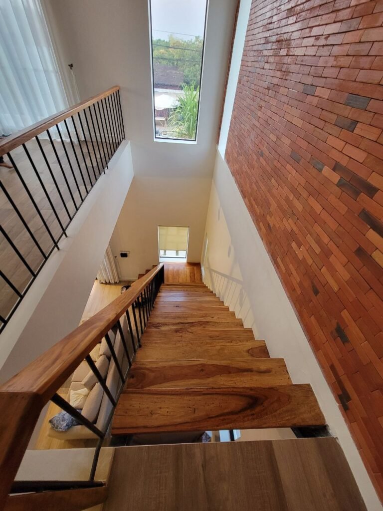 View looking down a modern wooden staircase with metal railings and a tall brick accent wall, large window at the landing, and natural light filling the space.