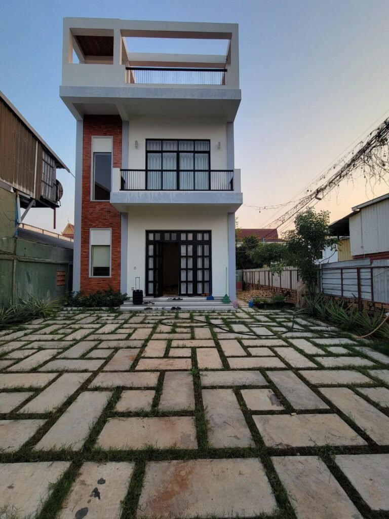 A modern three-story house with large windows, a balcony, and a flat roof. The front yard features a geometric stone pathway with grass in between the stones, surrounded by a garden and neighboring houses.