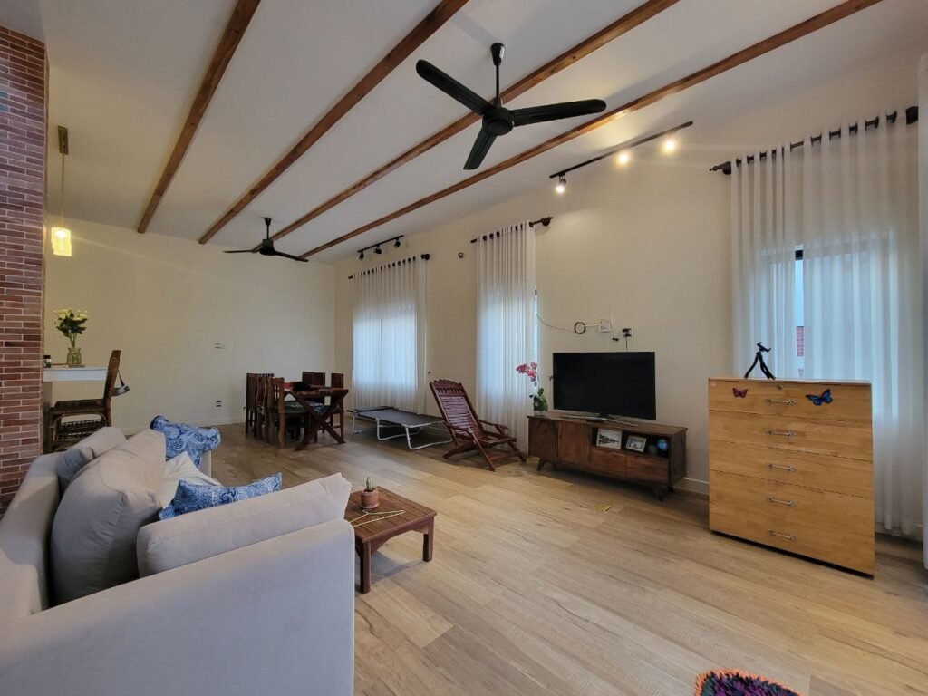 Spacious living room with light wood flooring, white walls, exposed ceiling beams, and large windows with sheer curtains. There’s a sofa, wooden dining table, TV on a console, fans, and a chest of drawers with butterfly decals.