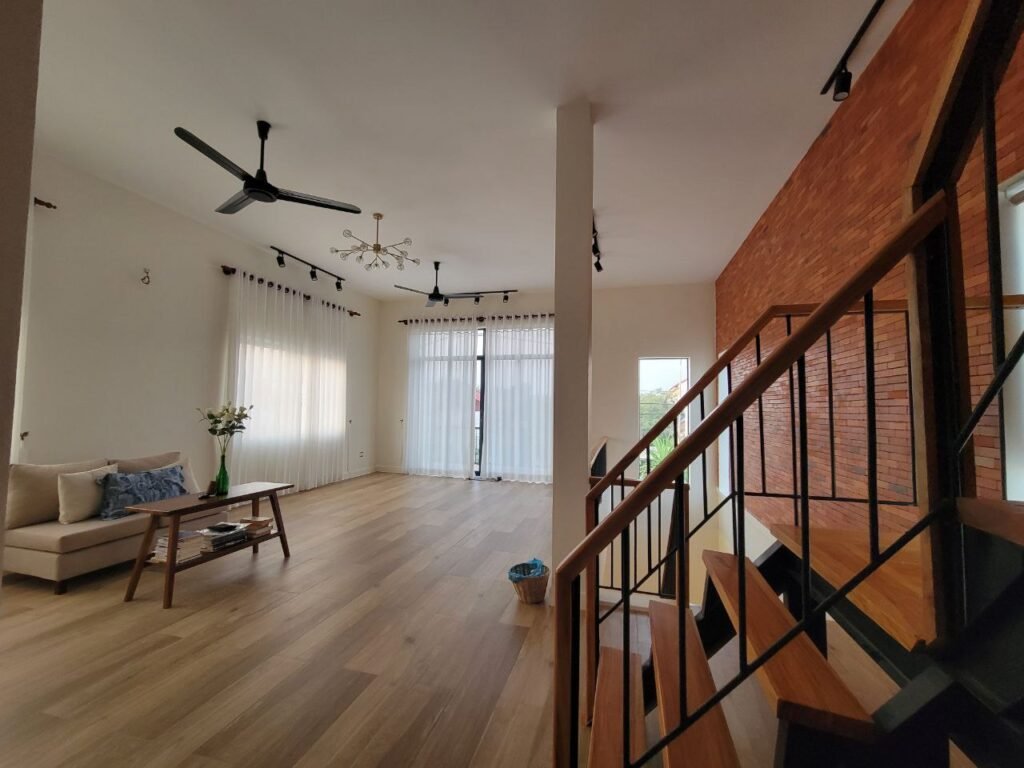 A modern living room with wooden floors, a beige sofa, large windows with sheer curtains, a brick accent wall, and a wooden staircase with black railings. Ceiling fans and track lighting are visible.