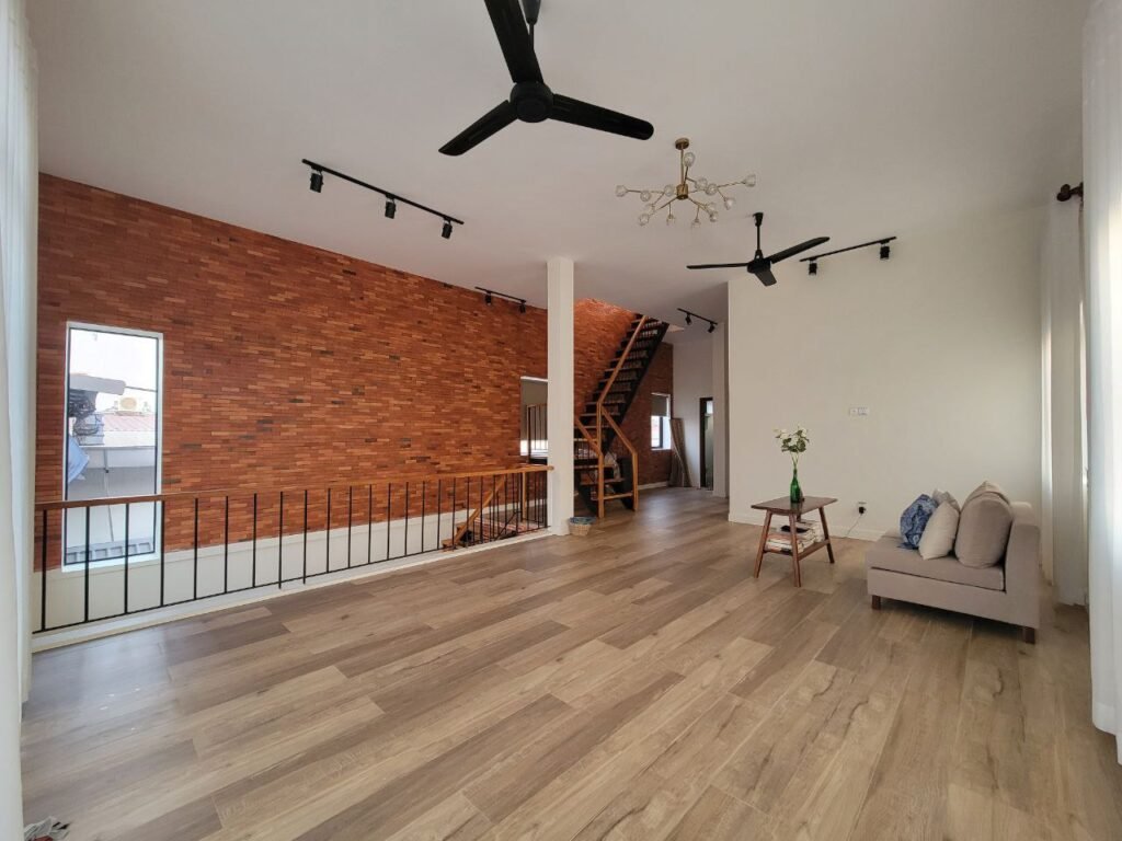 Spacious living area with light wooden floors, exposed brick accent wall, black ceiling fans, and a small couch with a coffee table. Staircase leads to an upper level, and large windows let in natural light.