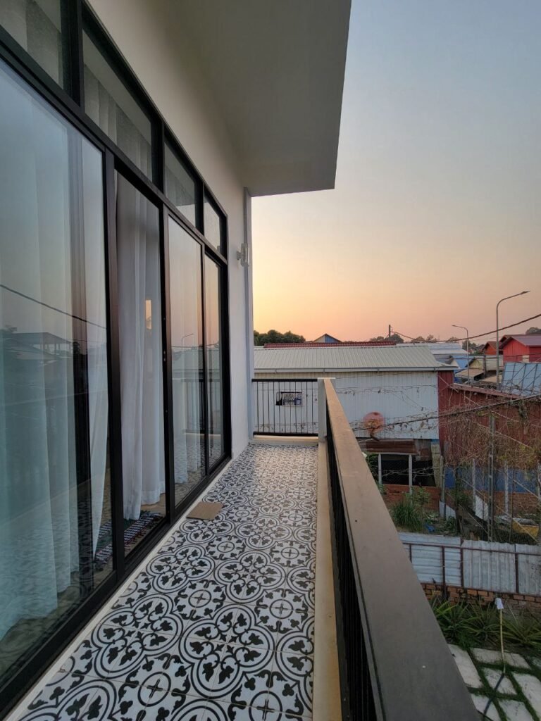 A patterned tile balcony with glass doors and a black railing overlooks houses and trees at sunset, with a clear sky above.