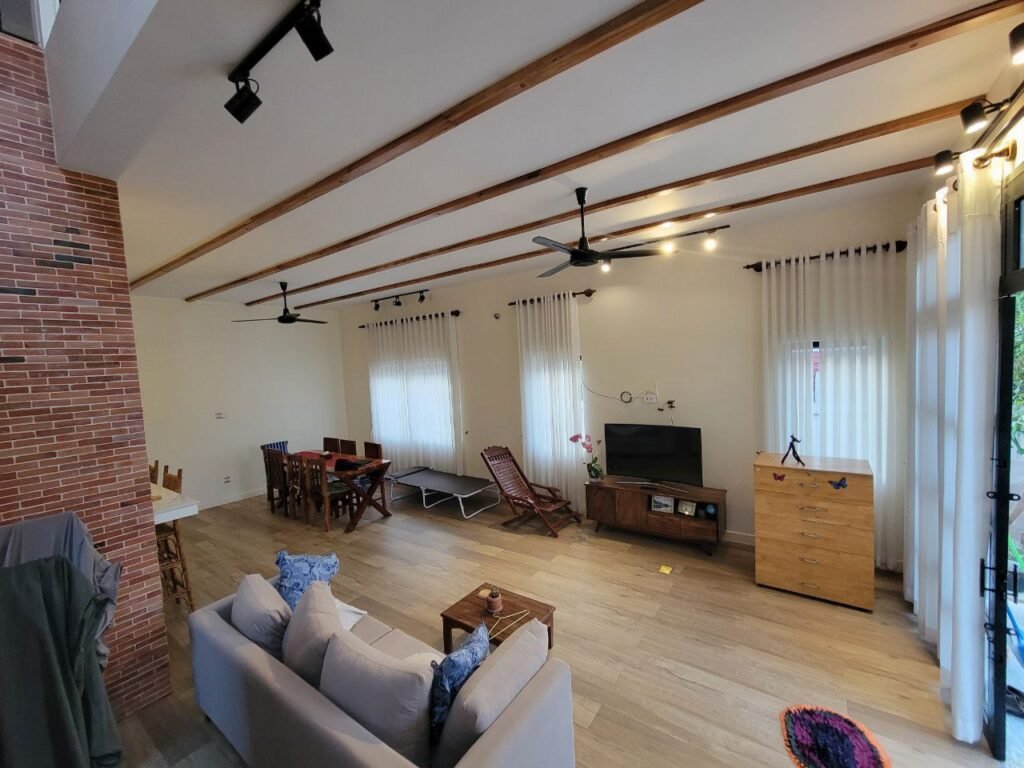 A spacious living room with wooden floors, exposed ceiling beams, and large windows with white curtains. The room features a sofa, TV, dining table, lounge chairs, and a brick accent wall.