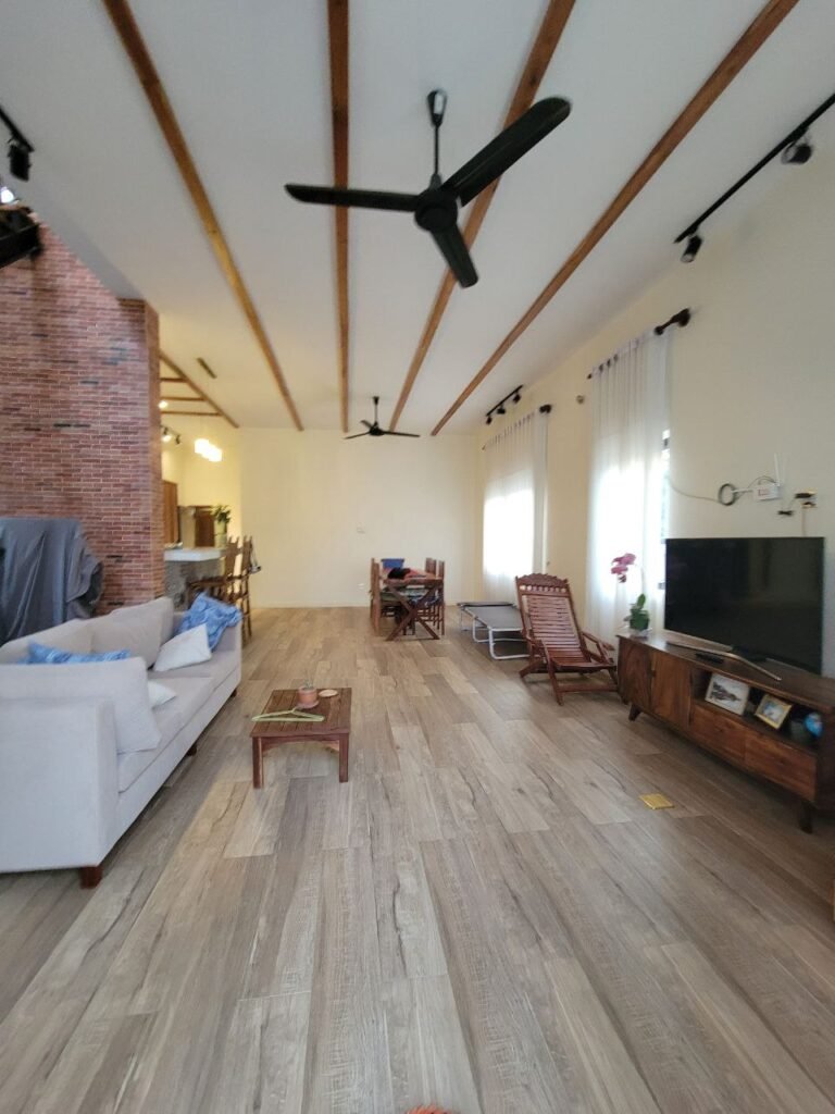 A spacious living room with wooden flooring, white walls, and exposed wooden ceiling beams. There are ceiling fans, a sofa, coffee table, TV, armchairs, potted plants, and large windows with sheer white curtains.