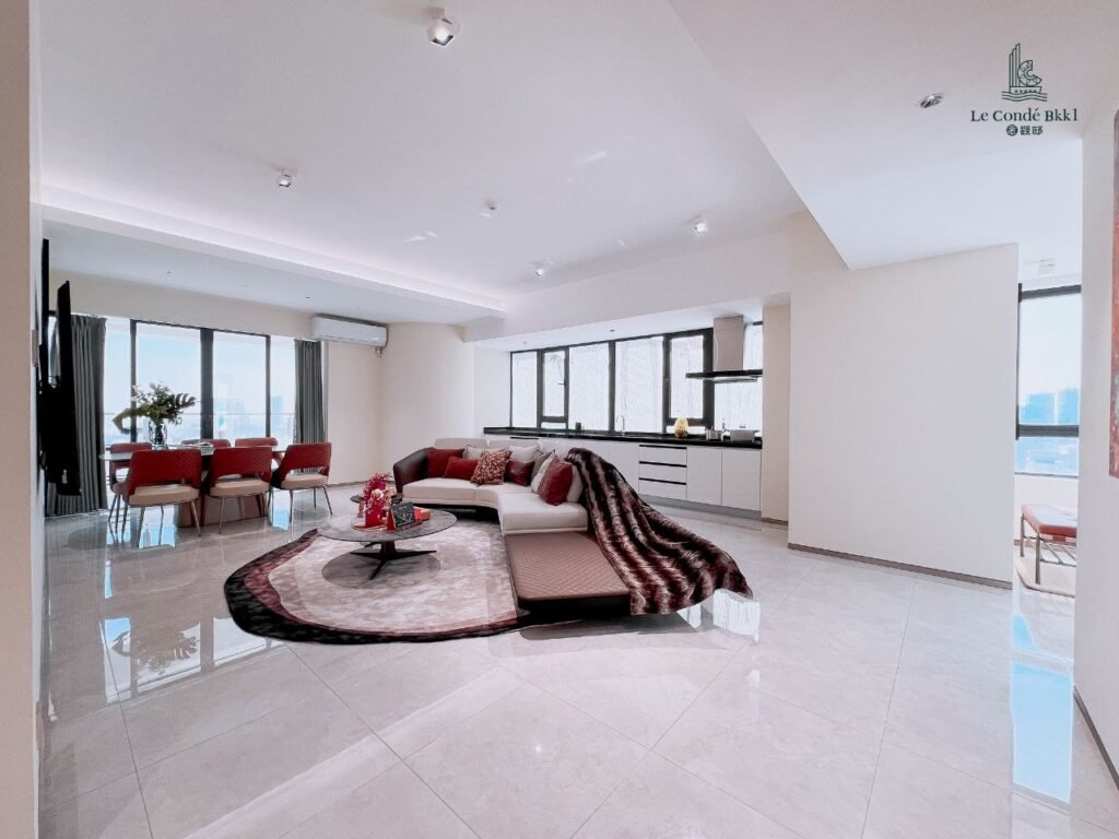 A spacious, modern studio living area with glossy tile floor, white walls, a red and white sectional sofa, a round rug, a dining table with red chairs, large windows, and an open kitchen.