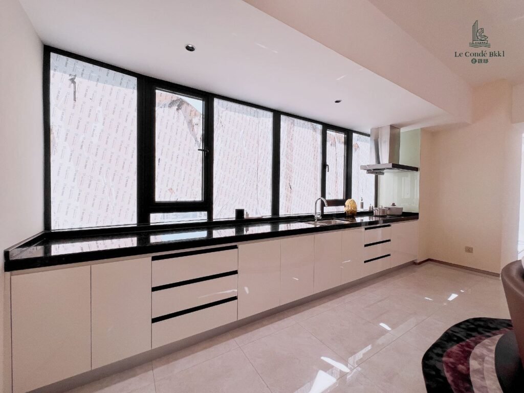 Modern studio kitchen with glossy white cabinets, black countertops, large windows letting in natural light, a built-in sink, stove, and range hood. Light-colored floor tiles and minimal decor create a clean, spacious feel.