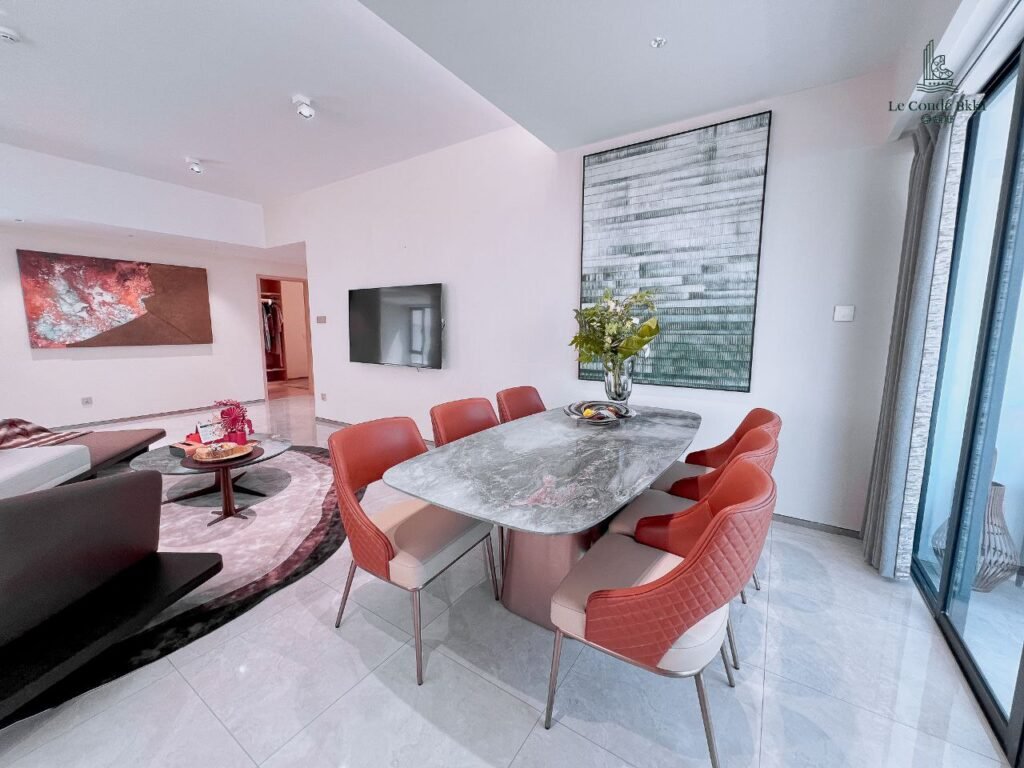 Modern dining area with a marble table, eight orange chairs, a vase of flowers, and large artwork. Adjacent studio-style living room with a circular rug, sofas, and wall art. Light-filled space with floor-to-ceiling windows.
