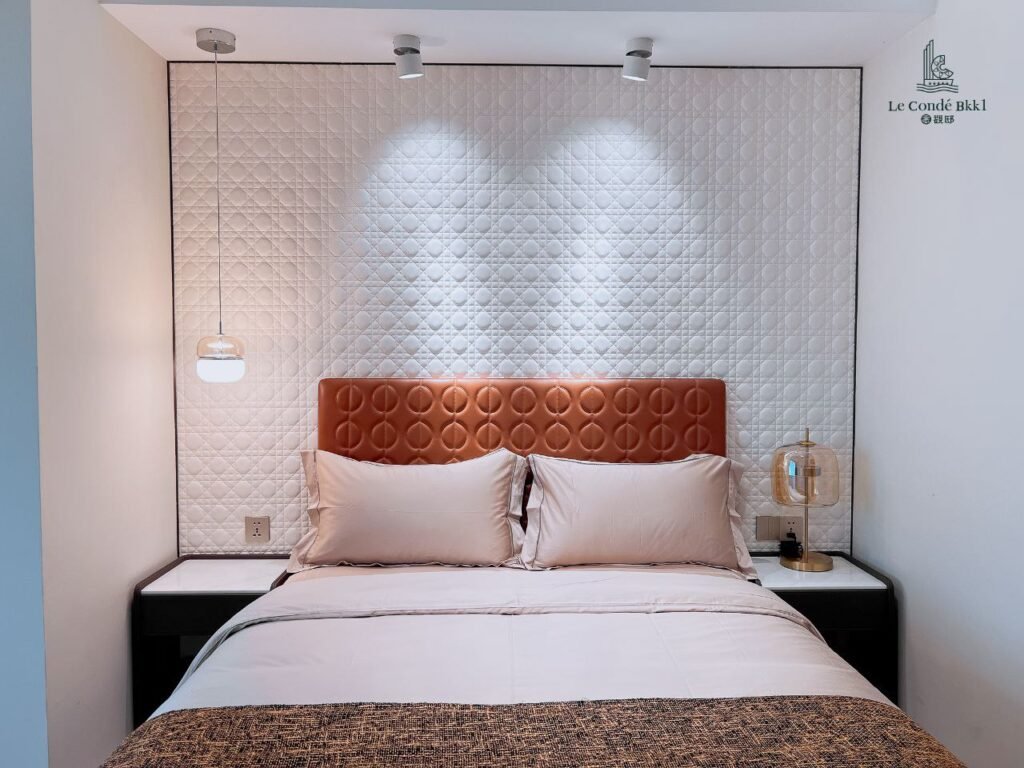 A neatly made bed with light beige bedding and a tufted brown headboard anchors this stylish studio, flanked by two bedside tables with modern lamps, set against a textured white accent wall under two ceiling spotlights.