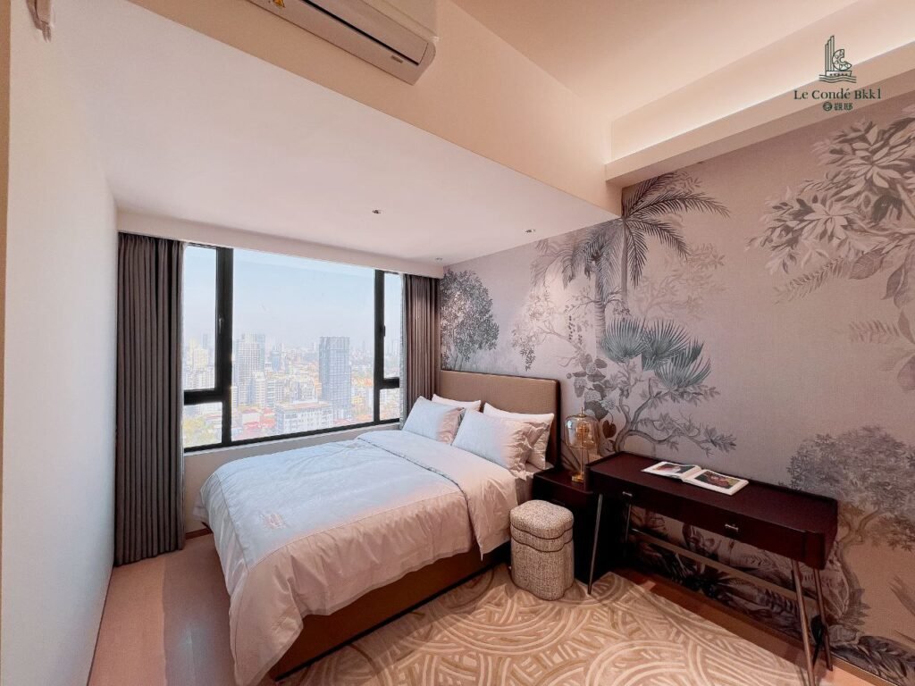 A cozy studio bedroom with a large window showing a city view, a double bed with white bedding, a patterned accent wall, a desk with chair, and neutral decor tones.