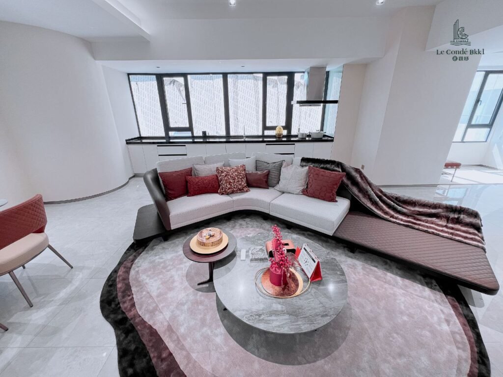 Modern studio living room with a curved sectional sofa, plush throw, accent pillows, round coffee table with decor, and large windows letting in natural light. The space features soft neutral and pink tones with contemporary styling.