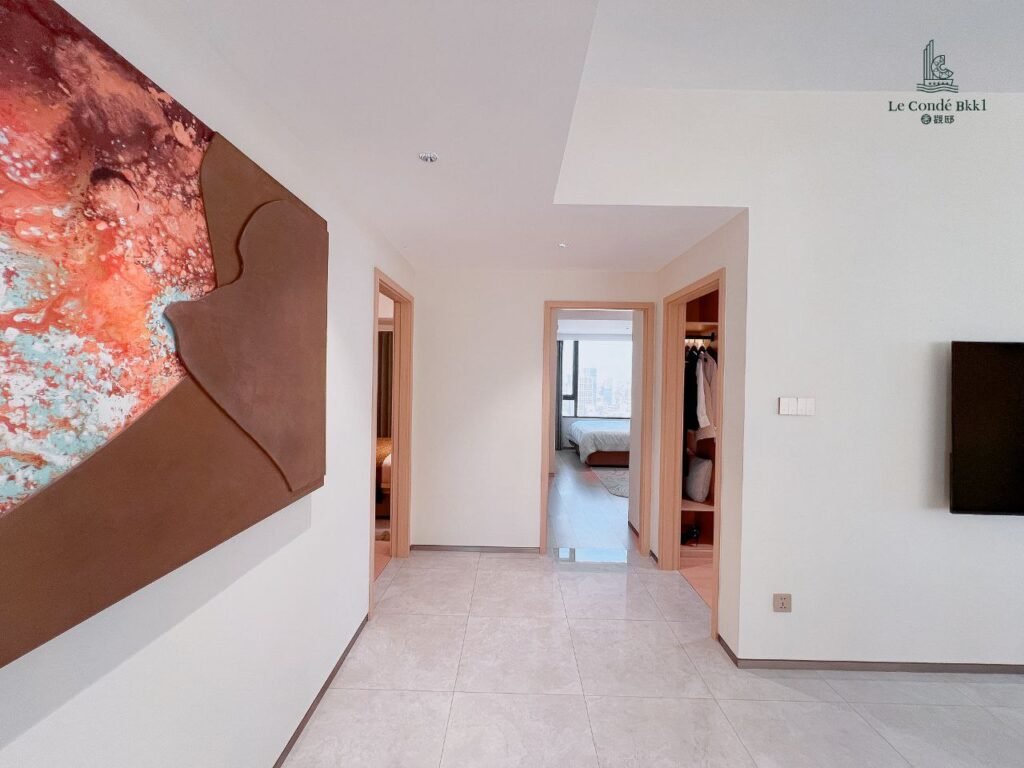 A bright, modern hallway with tiled floors and abstract wall art on the left leads into a studio bedroom with a large window and closet. Open doorways reveal airy spaces, while a mounted TV enhances the right wall.