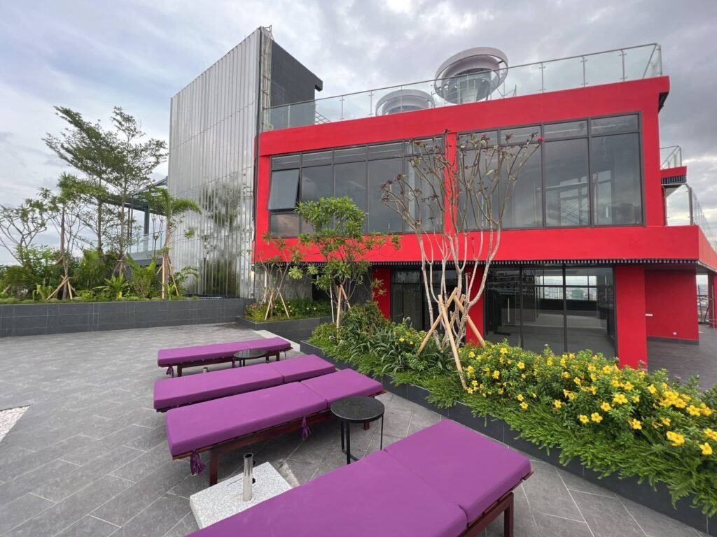 A modern red building with large glass windows sits at Time Square 3, surrounded by greenery and yellow flowers. In the foreground, purple cushioned benches and small round tables rest on a gray tiled patio under a cloudy sky.