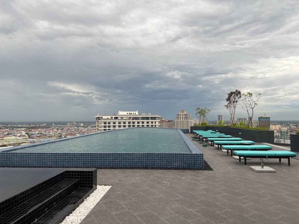 A rooftop infinity pool with turquoise lounge chairs lines one side, overlooking a cityscape near Time Square 3 under a cloudy sky. Some small trees are planted nearby, creating a modern, relaxing atmosphere.