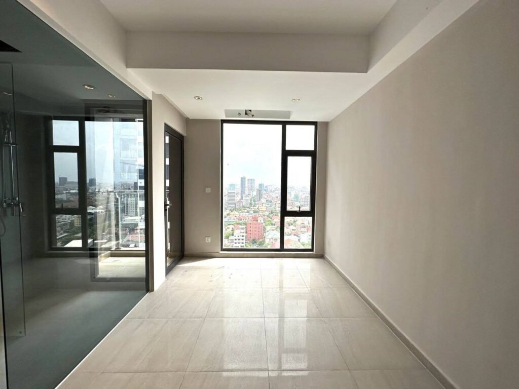 A bright, empty room with large floor-to-ceiling windows overlooking a cityscape, just steps from Time Square 3. The room features light-colored tiled floors, off-white walls, and a glass-walled shower area on the left.