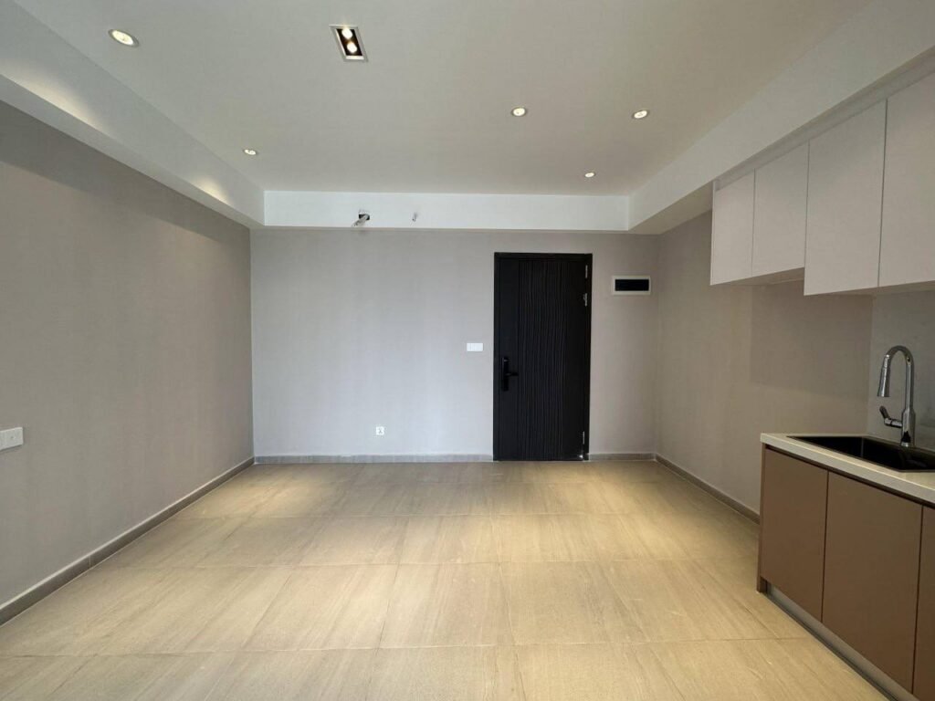 Minimalist, empty Time Square 3 room with a light beige tiled floor, gray walls, recessed ceiling lights, a closed black door, and a kitchen area with cabinets and sink on the right side.